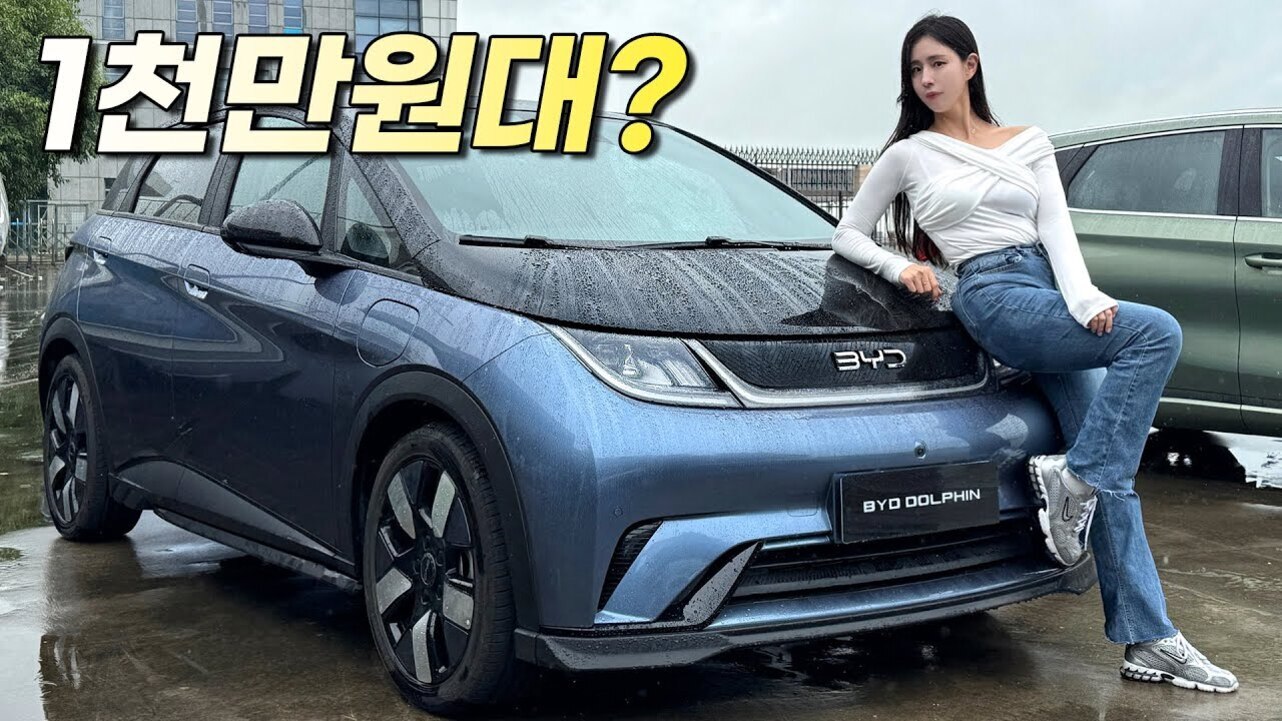 비 오는 날 파란색 BYD DOLPHIN 자동차 앞에 흰색 상의와 청바지를 입은 여성이 앉아 있고, 상단에는 '1천만원대?'라는 노란 글씨가 있다.