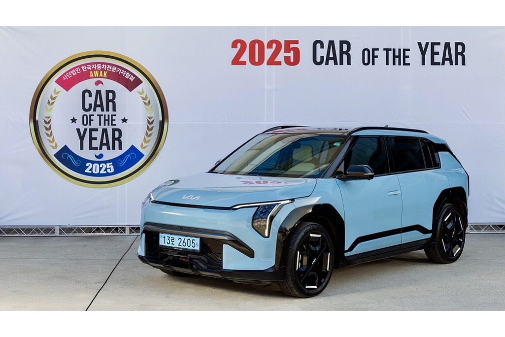 2025 올해의 차로 선정된 푸른색 기아 SUV 차량이 행사장 배경 앞에 전시되어 있다. 왼쪽에는 'CAR OF THE YEAR 2025'라고 적힌 원형 엠블럼이 보인다.