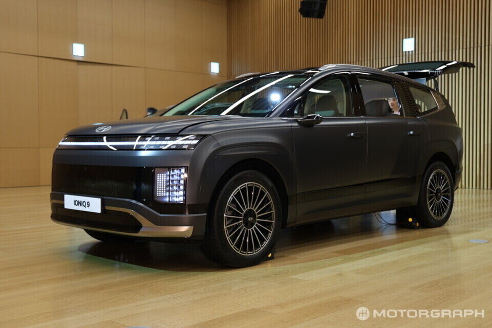 실내 전시장에 전시된 현대 아이오닉 9 SUV 차량이 보인다. 차량은 검정색이며, 뒷문과 트렁크가 열려 있다.