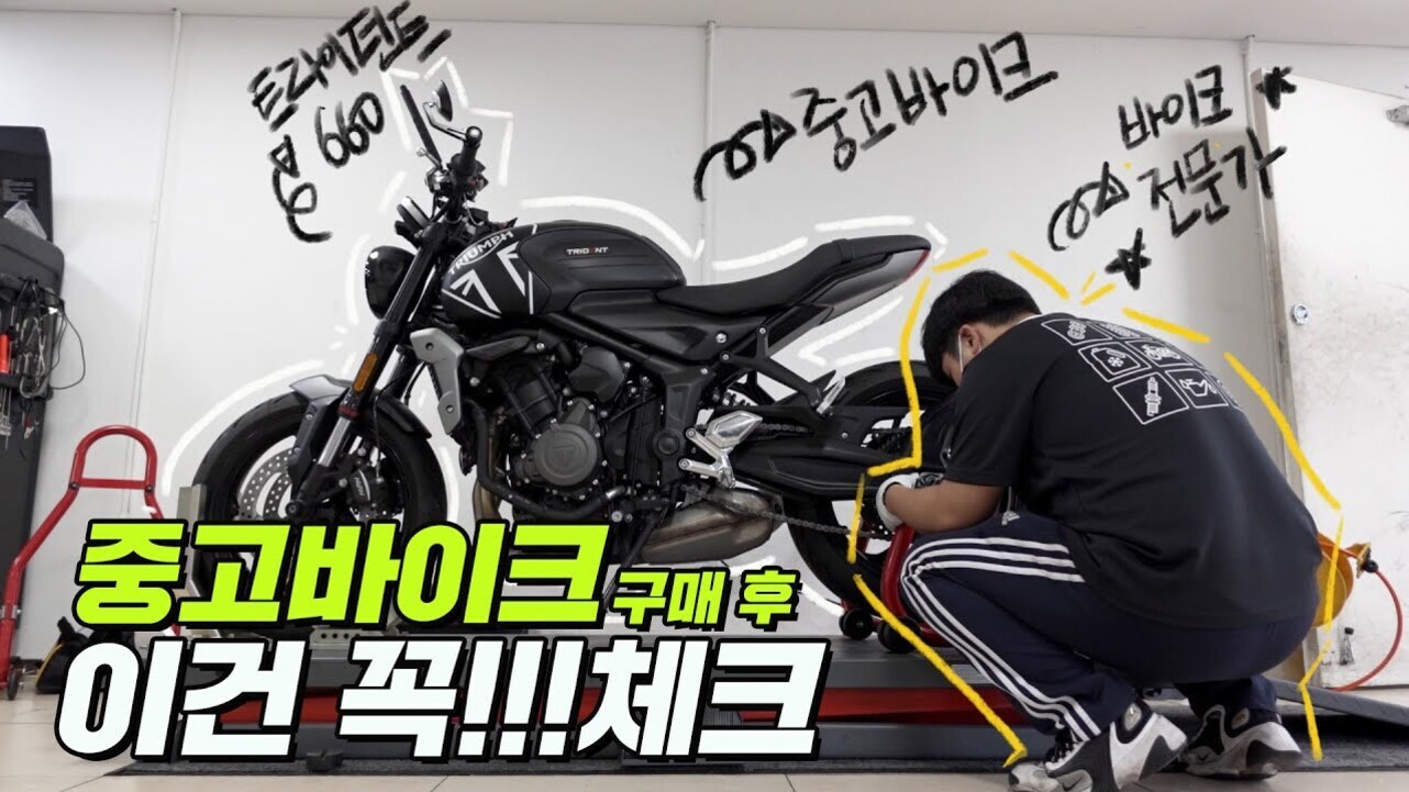 정비소에서 한 남성이 트라이엄프 트라이던트 660 중고 바이크를 점검하고 있는 모습. 벽에는 '중고바이크', '전문가' 등의 문구와 노란색 강조선이 그려져 있으며, 하단에 '중고바이크 구매 후 이건 꼭!! 체크'라는 큰 글자가 있다.