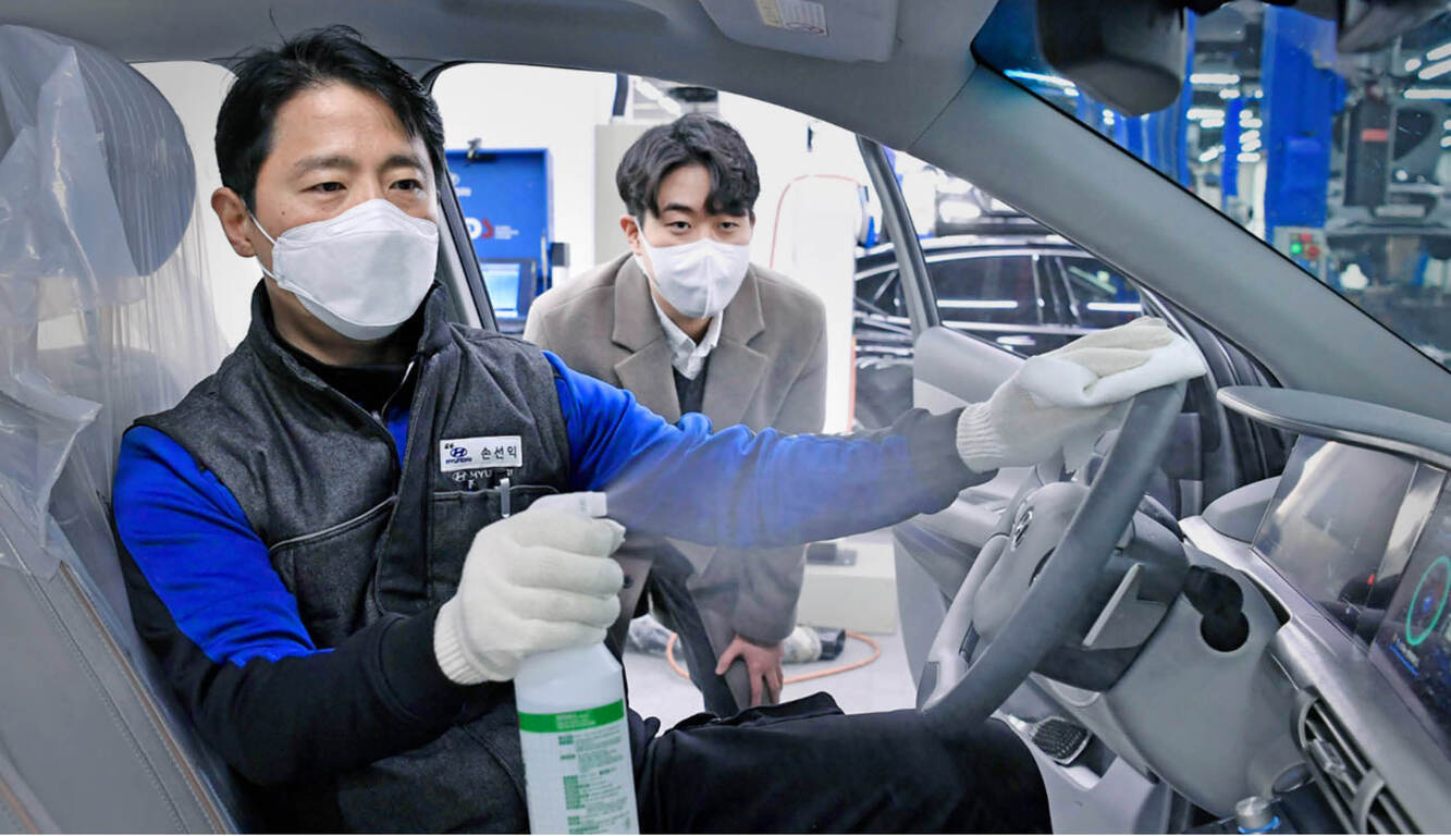 마스크를 쓴 남성이 자동차 내부에서 소독제를 분사하며 운전대를 닦고 있고, 다른 한 명이 이를 지켜보고 있다.