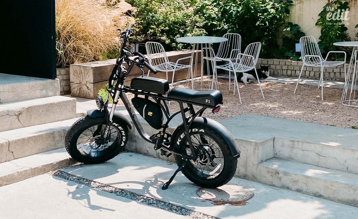 카페 야외 테라스에 전기 자전거 한 대가 세워져 있고, 뒤쪽에는 흰색 철제 의자와 테이블, 검은색과 흰색 고양이가 있다.