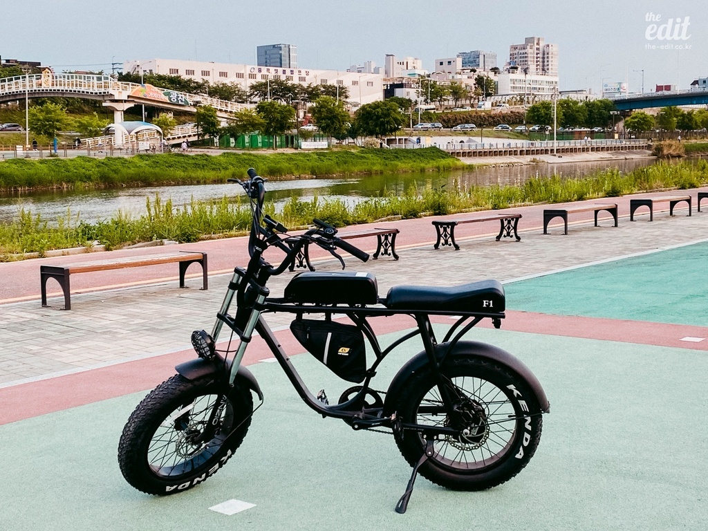 강변 산책로 옆에 전기 자전거 한 대가 세워져 있고, 주변에는 벤치와 강, 그리고 도시 건물들이 보인다.