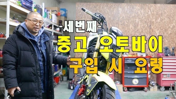 한 남성이 작업장 안에서 노란색 오토바이 옆에 서 있고, 화면에는 '세번째 중고 오토바이 구입 시 요령'이라는 큰 글자가 표시되어 있다.