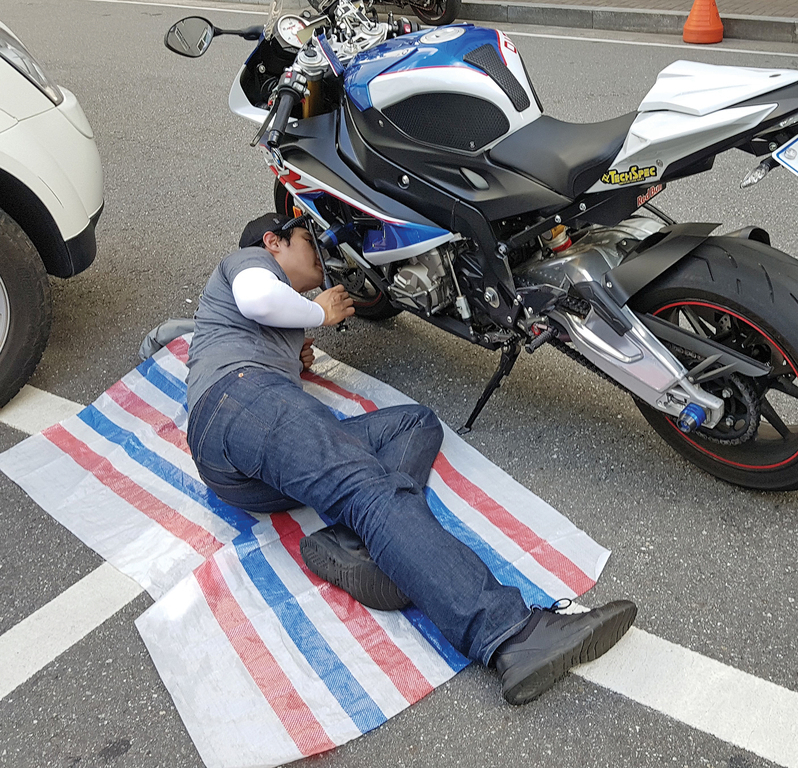 한 남성이 도로 위에 깔린 줄무늬 천 위에서 오토바이 아래를 점검하고 있다.
