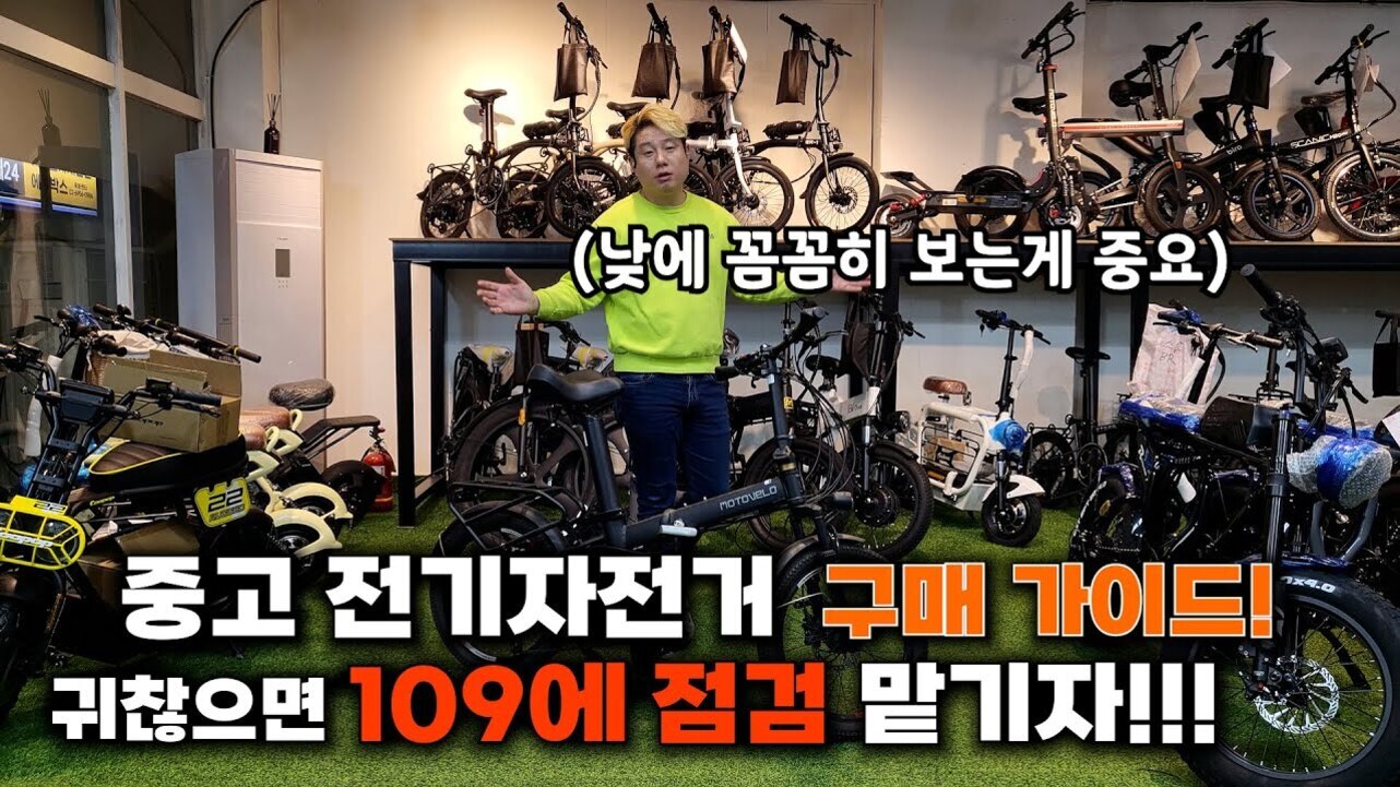 여러 대의 전기자전거가 진열된 매장에서 한 남성이 밝은 연두색 상의를 입고 서 있다. 화면 중앙에는 '(낮에 꼼꼼히 보는게 중요)'라는 자막이 있고, 하단에는 '중고 전기자전거 구매 가이드! 귀찮으면 109에 점검 맡기자!!!'라는 큰 글씨가 적혀 있다.